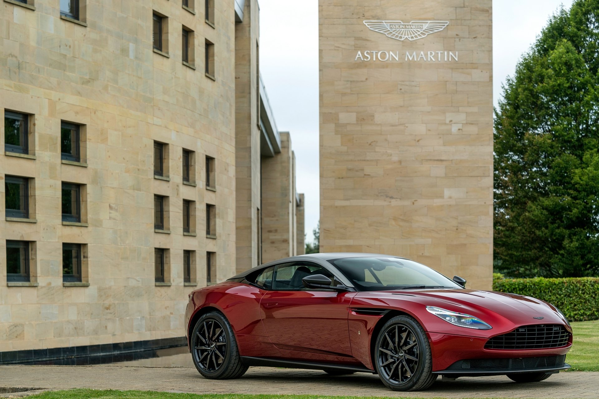 Red Aston Martin DB11 grand tourer supercar parked beside a sandstone building and trees — sleek vehicle shot rendered as a 4K Ultra HD PC desktop wallpaper and background.