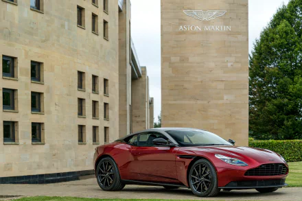 Red Aston Martin DB11 grand tourer supercar parked beside a sandstone building and trees — sleek vehicle shot rendered as a 4K Ultra HD PC desktop wallpaper and background.