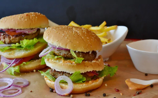HD PC desktop wallpaper showing two juicy hamburgers with lettuce, cheese and onion rings, served with fries — a lunch meal burger food background.