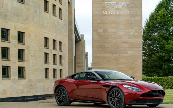 Red Aston Martin DB11 grand tourer supercar parked beside a sandstone building and trees — sleek vehicle shot rendered as a 4K Ultra HD PC desktop wallpaper and background.