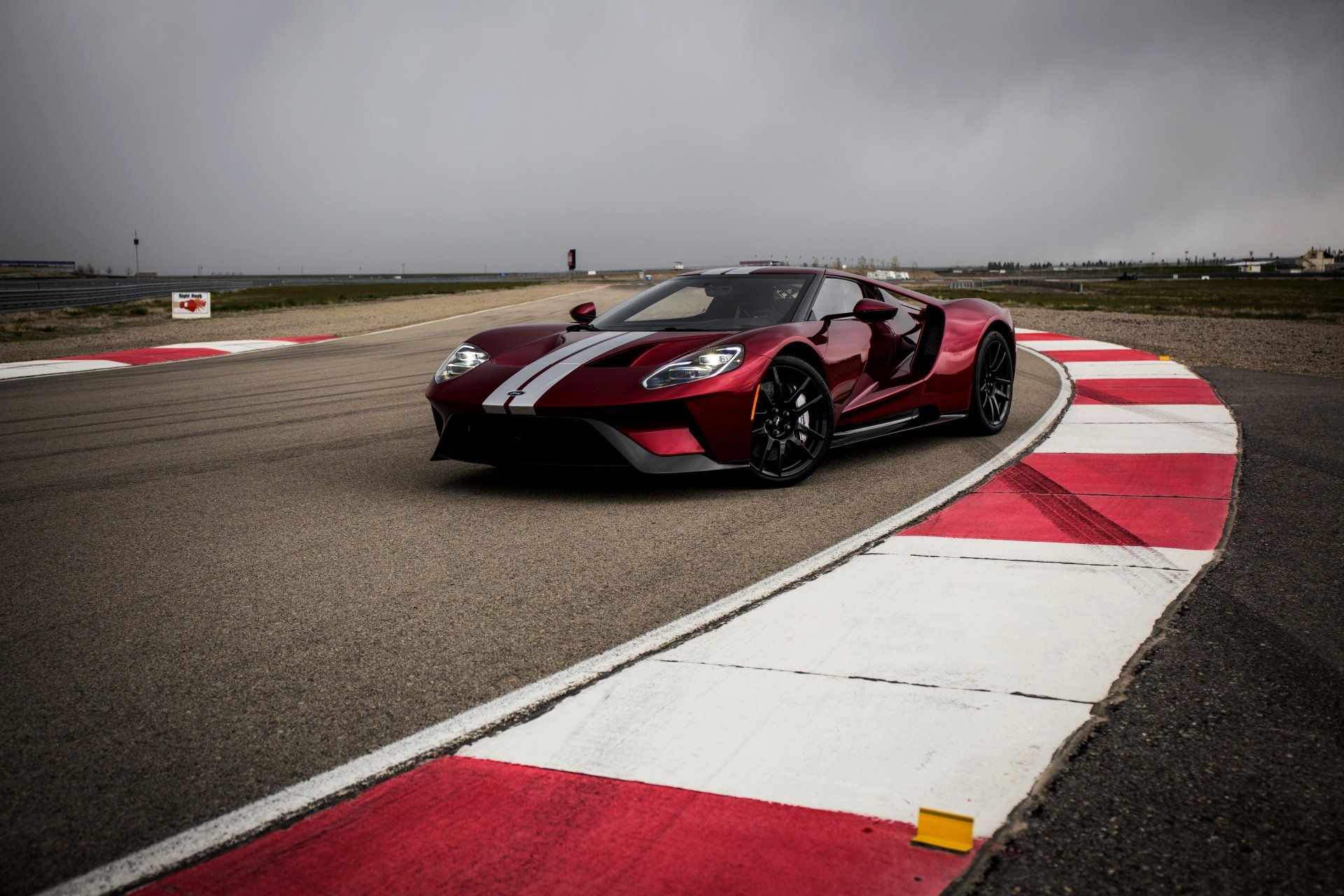 A sleek Ford GT supercar in deep red with white racing stripes speeds around a racetrack under a cloudy sky, captured in stunning 4K Ultra HD quality.