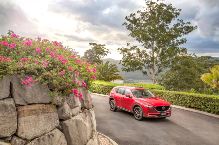 A red Mazda CX-5 SUV drives along a winding road surrounded by lush greenery and vibrant pink flowers, captured in a 4K Ultra HD desktop wallpaper.