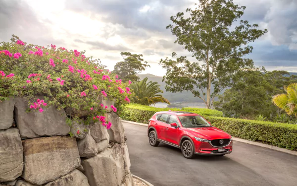 A red Mazda CX-5 SUV drives along a winding road surrounded by lush greenery and vibrant pink flowers, captured in a 4K Ultra HD desktop wallpaper.