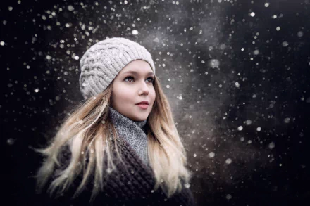 HD PC desktop wallpaper of a blonde woman model in a knit hat and scarf, hazel eyes gazing upward as snowflakes drift around her.