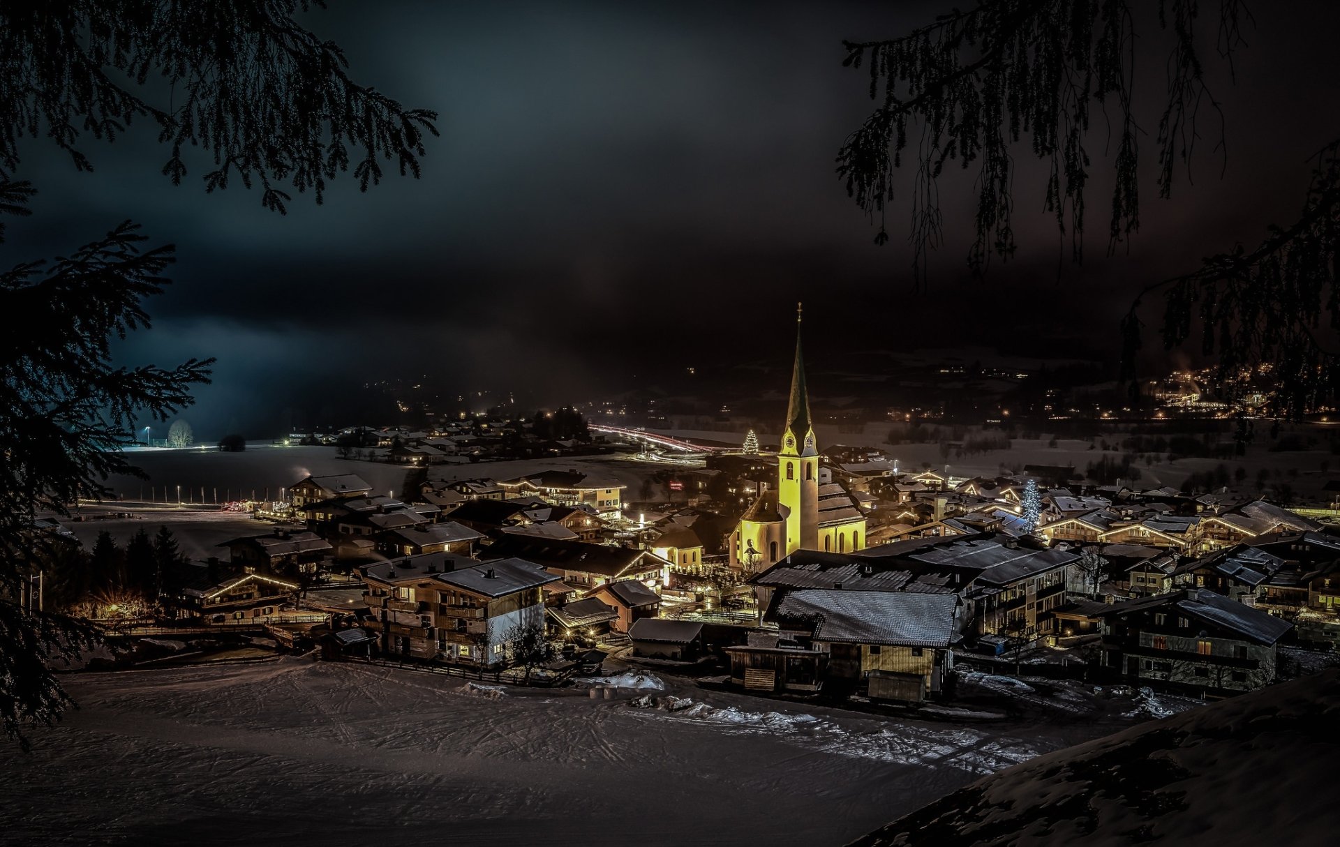 A picturesque winter night in an Austrian village, featuring a beautifully illuminated church surrounded by snow-covered rooftops under a dark sky.