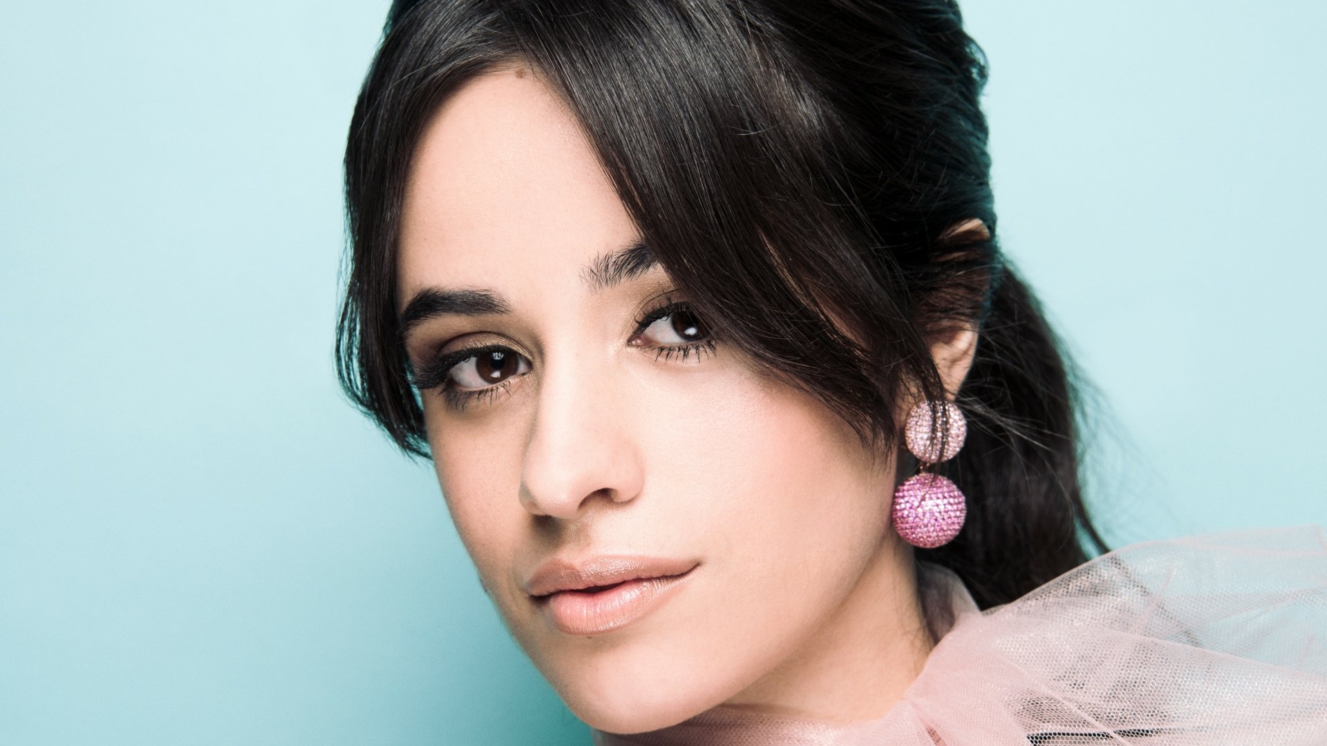 Close-up 4K Ultra HD portrait of a Latina singer with brunette hair, brown eyes, and sparkling earrings, set against a soft blue background.