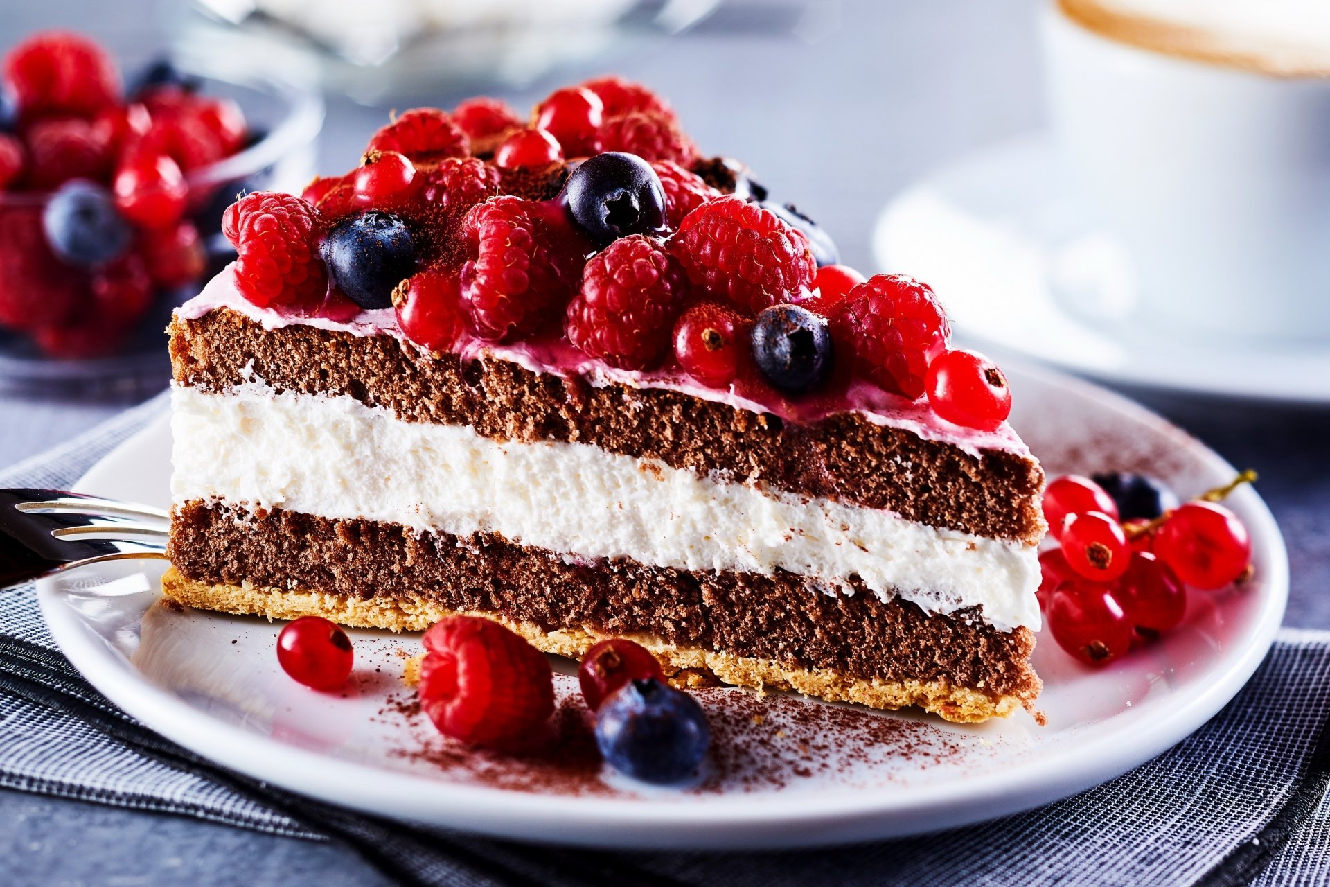A slice of chocolate cake layered with cream and topped with fresh blueberries, raspberries, and currants on a white plate, set against a blurred background.