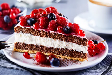 A slice of chocolate cake layered with cream and topped with fresh blueberries, raspberries, and currants on a white plate, set against a blurred background.