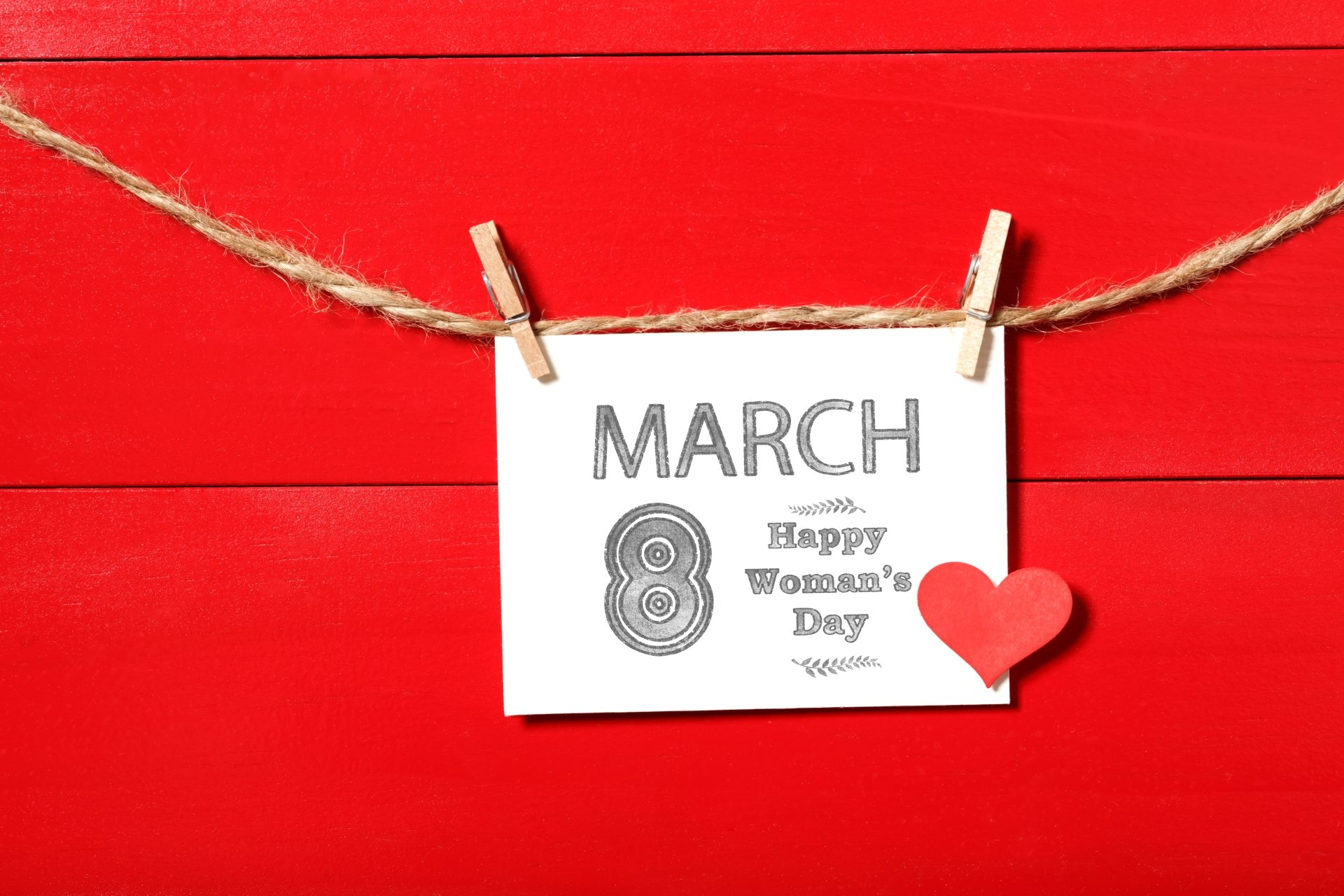 A 4K Ultra HD Women's Day wallpaper featuring a white card with March 8 Happy Women's Day clipped on a twine against a bright red wooden background with a small red heart.