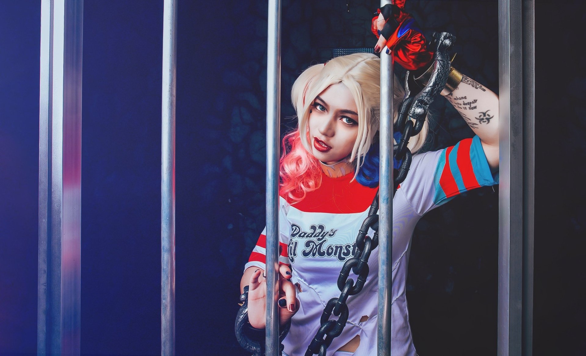 HD desktop wallpaper of a blonde woman cosplaying Harley Quinn with blue eyes, red lipstick, and tattoos, posing behind metal bars while holding a chain.