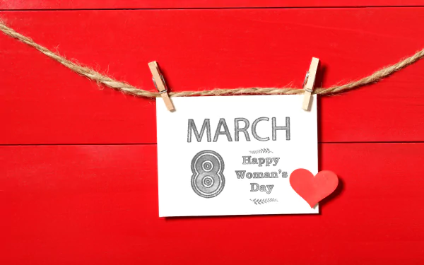 A 4K Ultra HD Women's Day wallpaper featuring a white card with March 8 Happy Women's Day clipped on a twine against a bright red wooden background with a small red heart.