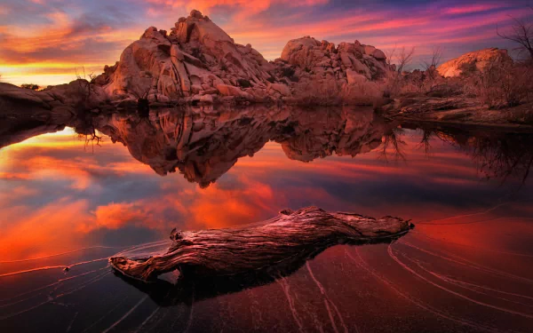 HD wallpaper of a serene lake at sunset, with driftwood floating on the water and vibrant reflections of the colorful sky and rocky landscape. Nature's beauty captured in a stunning desktop background.