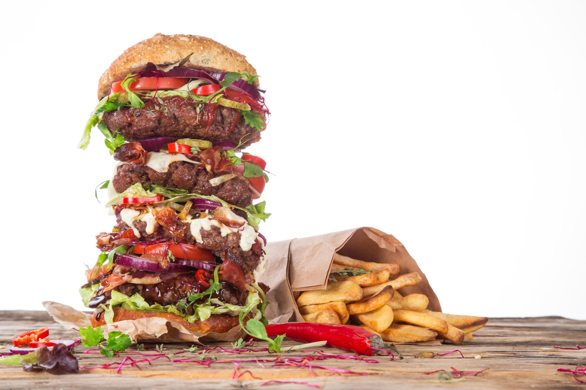 A 4K Ultra HD still life of a towering burger stacked with peppers, fresh greens, and sauces, accompanied by crispy French fries on a rustic wooden surface.