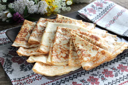 HD desktop wallpaper featuring a plate of neatly folded crêpes on a traditional embroidered cloth, with white flowers in the background.