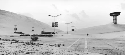 Black-and-white Mad Max video game 5K Ultra HD PC desktop wallpaper showing a deserted parking lot with wrecked cars, lampposts and a lone figure walking toward distant dunes.