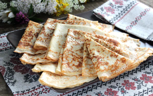 HD desktop wallpaper featuring a plate of neatly folded crêpes on a traditional embroidered cloth, with white flowers in the background.