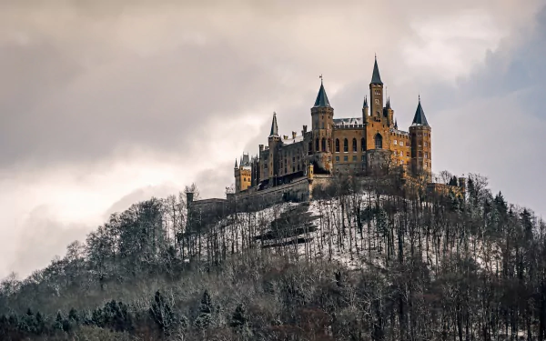 HD wallpaper of Hohenzollern Castle in Germany, perched atop a forested hill under a cloudy sky. The historic building stands majestically, showcasing its stunning architecture and serene surroundings.
