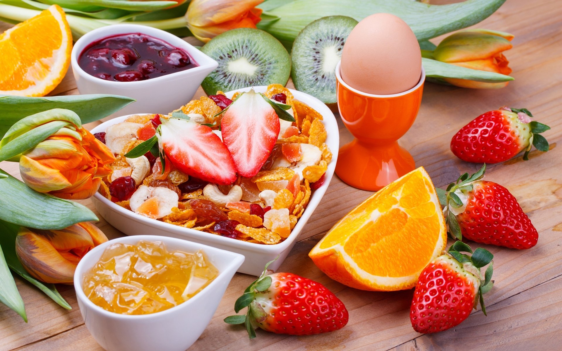 HD desktop wallpaper showing a colorful breakfast still life with jam, cereal topped with strawberries, a boiled egg, fresh fruit, and flower decorations on a wooden surface.