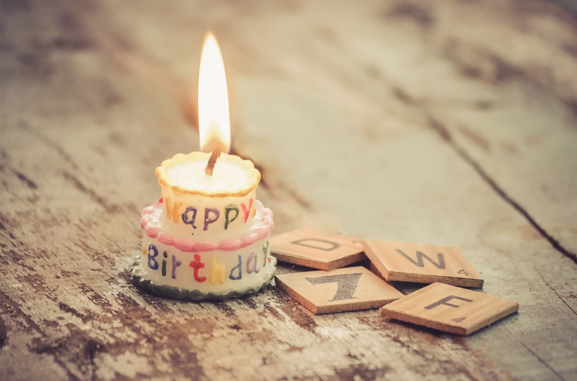 A lit candle shaped like a birthday cake with Happy Birthday text, surrounded by letter tiles on a wooden surface, captured in HD for a festive holiday mood.