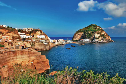 HD PC desktop wallpaper photograph of Ischia coastline: sunlit rocky cliffs and seaside houses, a small island rising from the blue ocean with a clear horizon.