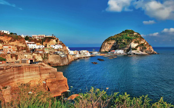 HD PC desktop wallpaper photograph of Ischia coastline: sunlit rocky cliffs and seaside houses, a small island rising from the blue ocean with a clear horizon.