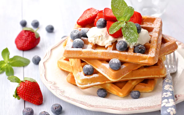 HD desktop wallpaper of a breakfast featuring golden waffles topped with fresh blueberries, strawberries, whipped cream, and mint leaves on a decorative plate.