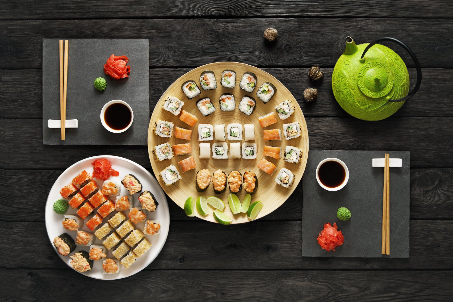 A 4K Ultra HD still life of sushi and seafood on plates with chopsticks, soy sauce, wasabi, and a green teapot arranged on a dark wooden table.