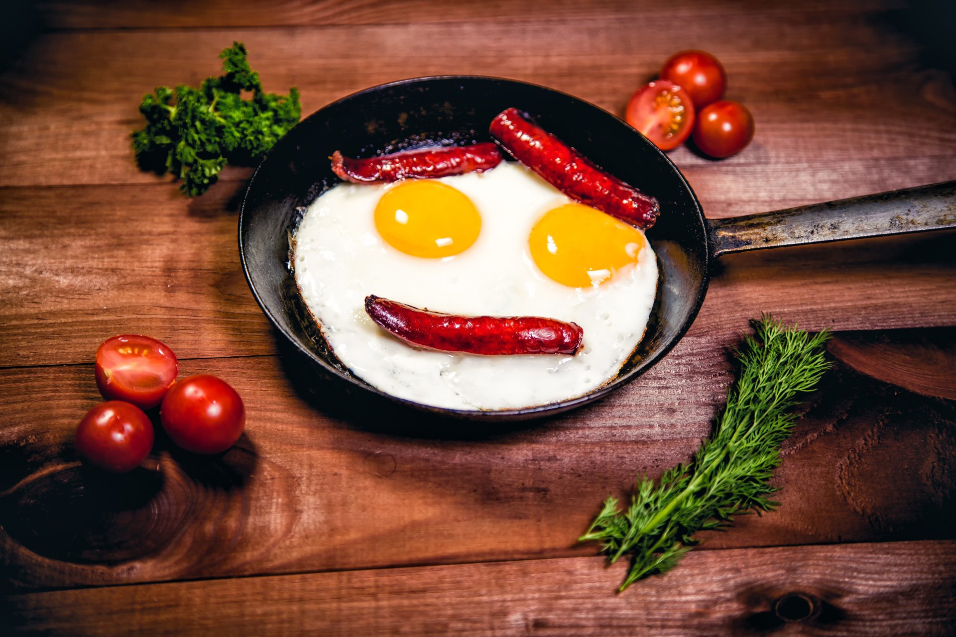 A 4K Ultra HD still life desktop wallpaper featuring a frying pan with two sunny-side-up eggs and sausages arranged to resemble a smiling face, surrounded by fresh tomatoes and herbs.