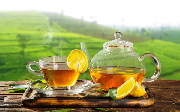 A 4K Ultra HD wallpaper featuring a clear glass teapot and cup of tea with lemon slices, set on a wooden tray against a blurred green hill background.