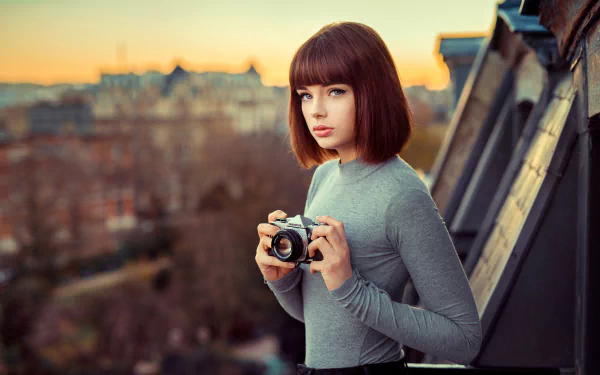 A brunette woman with short hair and blue eyes, holding an Olympus camera, captured in a depth of field style by Marie Grippon, set against a blurred cityscape at sunset.