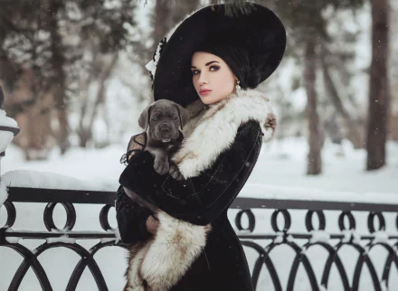 A woman in a black hat and fur coat holds a puppy in a snowy winter park, captured with a soft depth of field in this HD desktop wallpaper.