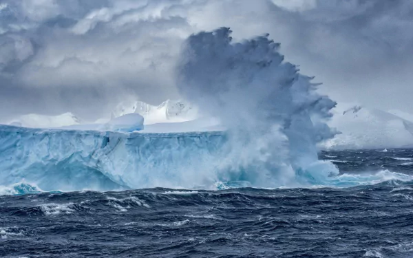 HD desktop wallpaper showcasing a powerful ocean wave crashing against a massive iceberg under a cloudy sky in a dramatic natural scene.