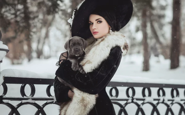 A woman in a black hat and fur coat holds a puppy in a snowy winter park, captured with a soft depth of field in this HD desktop wallpaper.