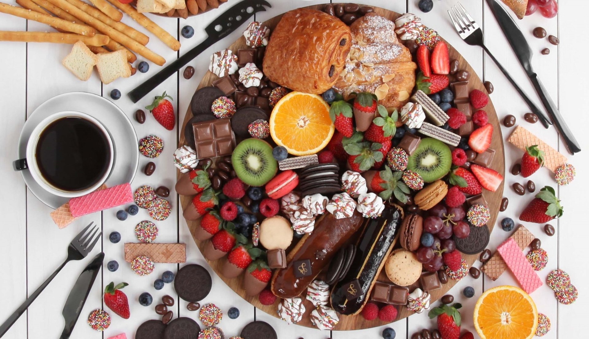 HD desktop wallpaper featuring a still life of viennoiserie, éclair, chocolate, fresh strawberries, kiwi, orange slices, assorted sweets, and a cup of coffee.