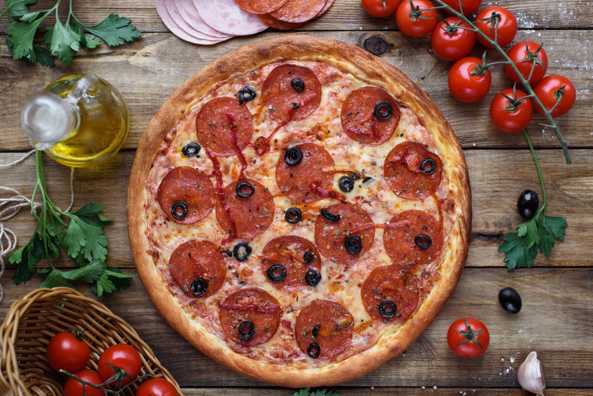 HD PC desktop wallpaper featuring a still life of a pepperoni pizza with olives, fresh tomatoes, olive oil, and herbs on a rustic wooden surface.