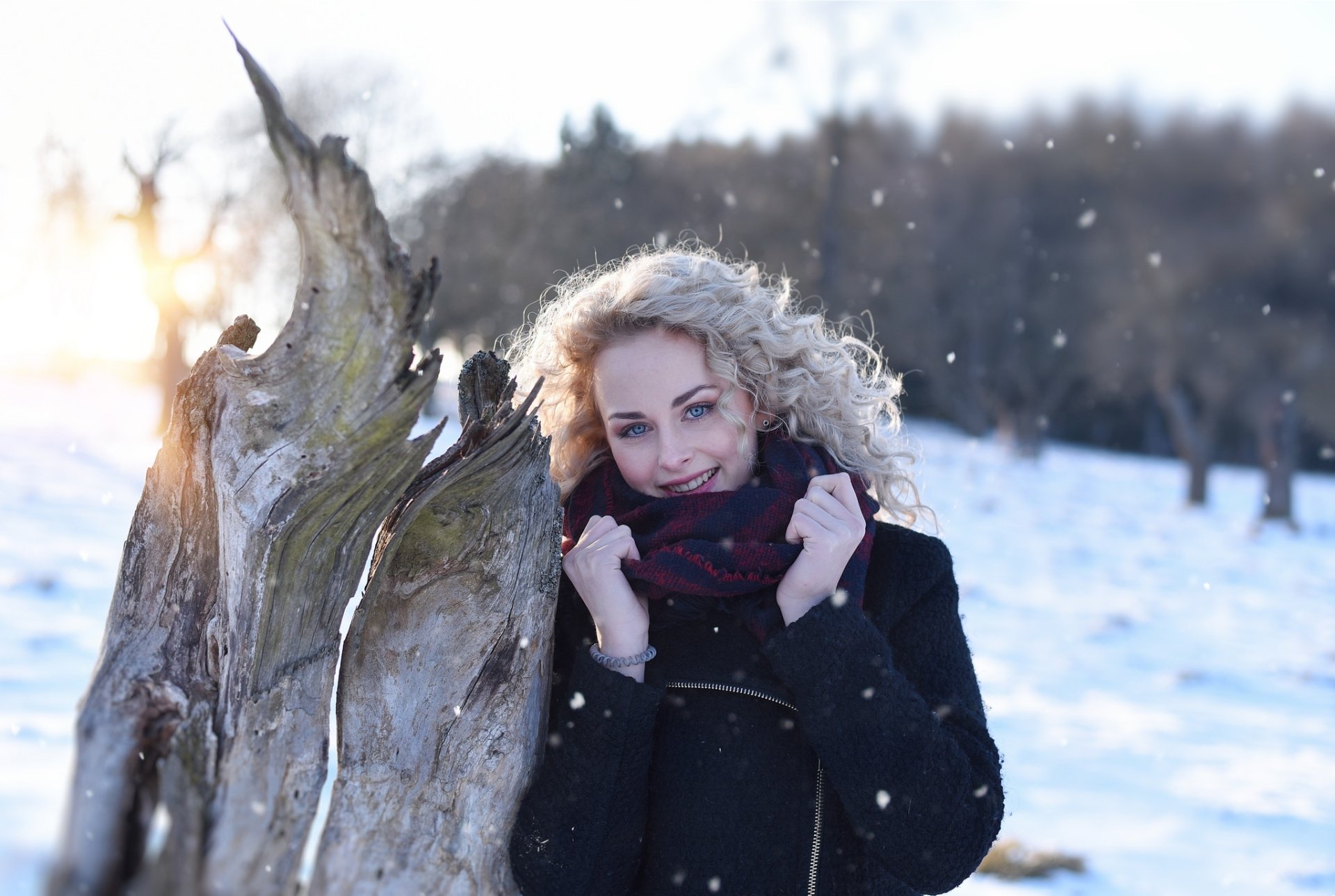 A smiling blonde woman with blue eyes and a scarf stands near a tree trunk in a snowy landscape, captured with a shallow depth of field in this HD desktop wallpaper.