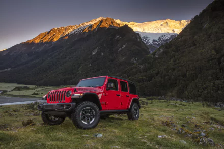 Red Jeep Wrangler off-road vehicle parked on grassy mountain meadow at sunrise, 2K Quad HD PC desktop wallpaper background.