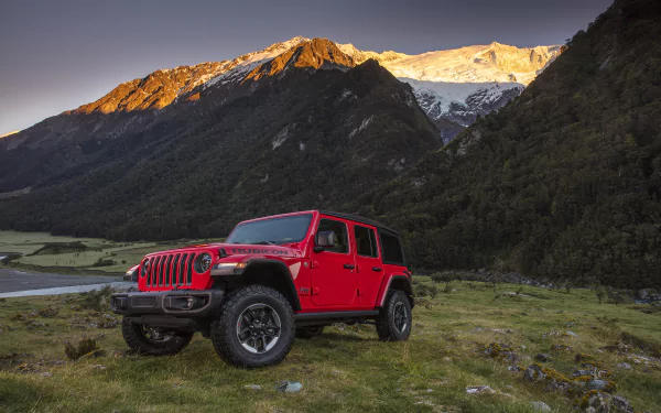 Red Jeep Wrangler off-road vehicle parked on grassy mountain meadow at sunrise, 2K Quad HD PC desktop wallpaper background.