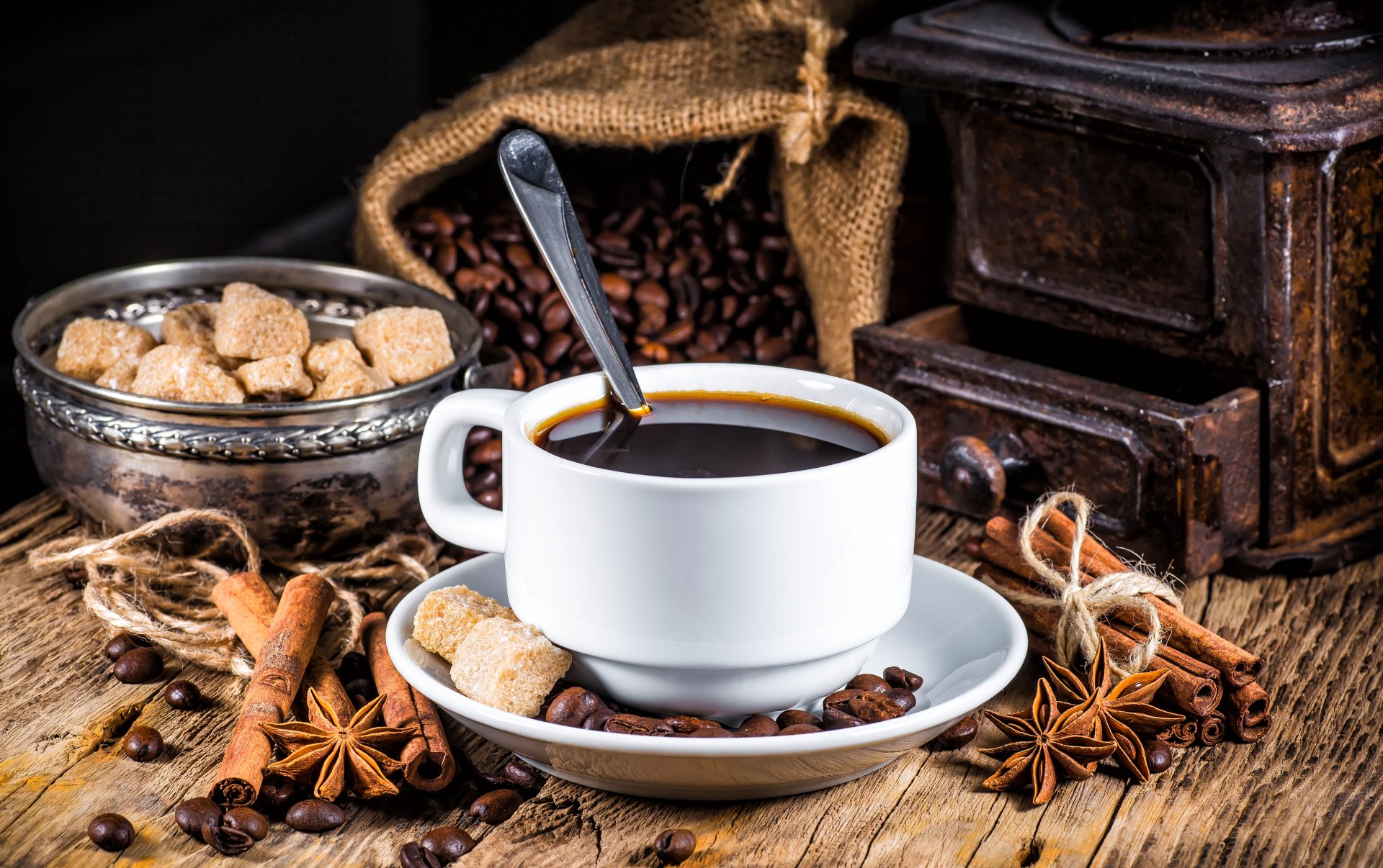 A 4K Ultra HD still life of a white coffee cup with a spoon, surrounded by cinnamon sticks, star anise, coffee beans, brown sugar cubes, and a vintage grinder on a wooden table.