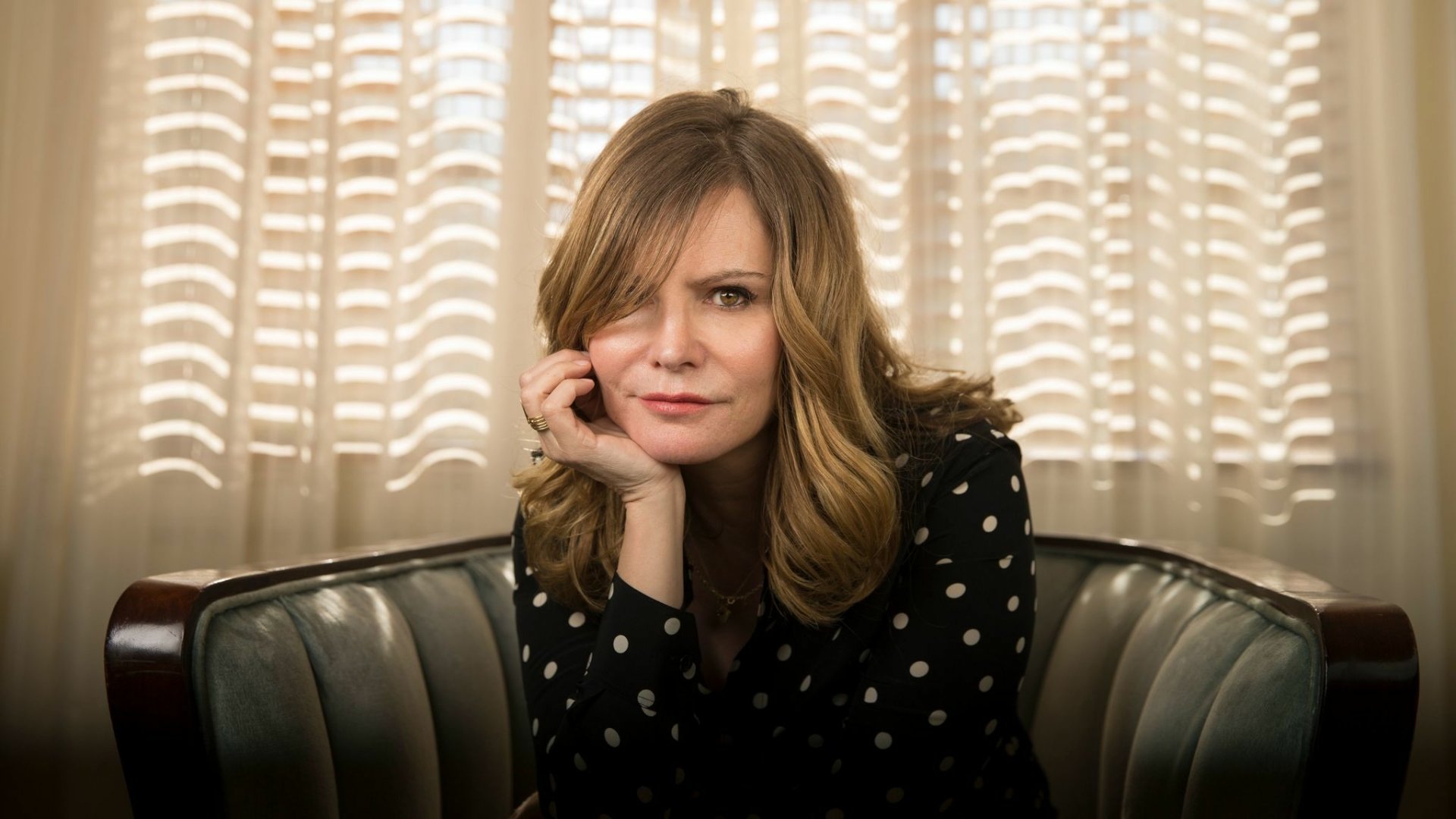 Jennifer Jason Leigh sitting thoughtfully in a booth, a high-definition wallpaper with a backlit window pattern background.