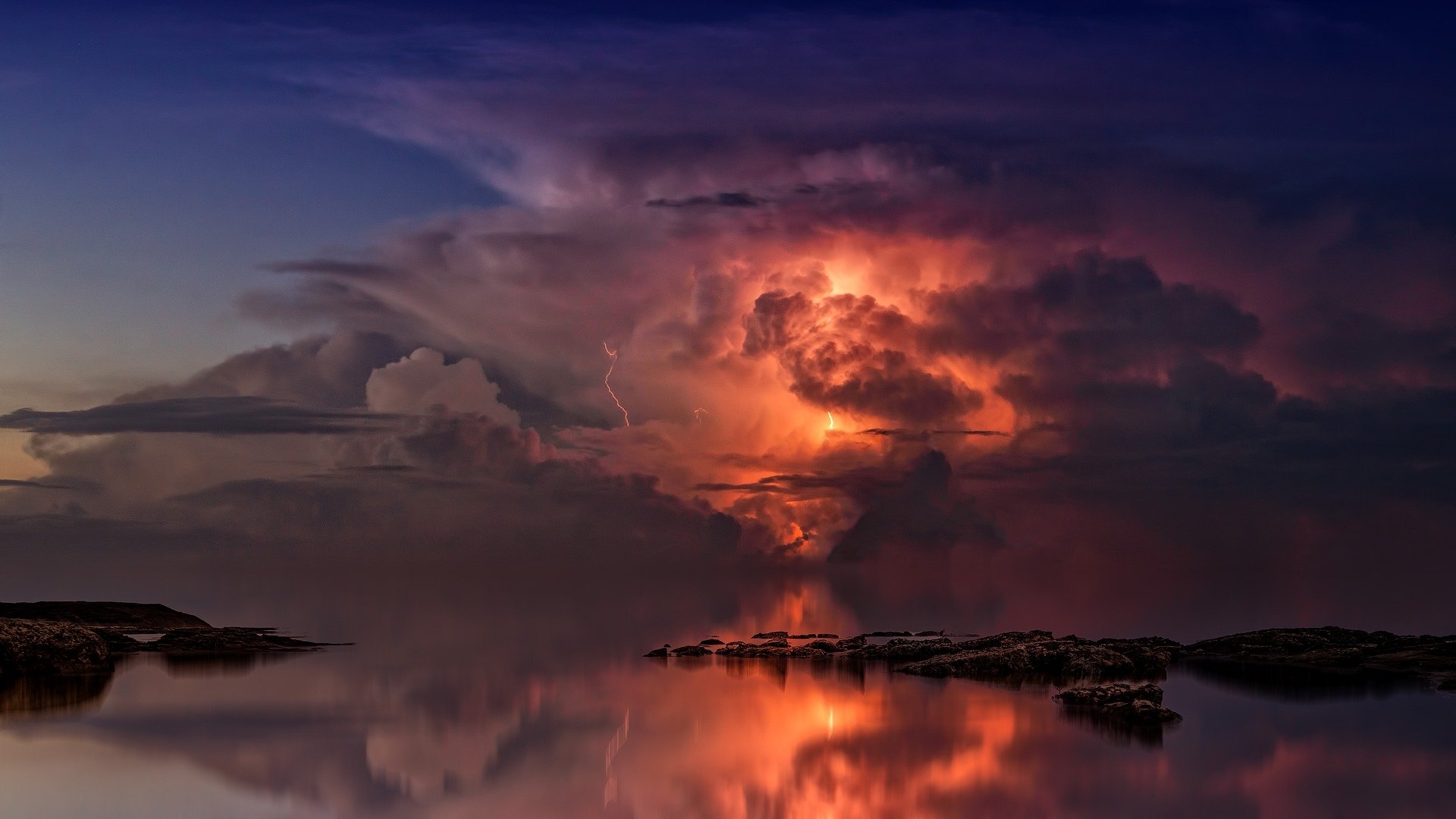 HD desktop wallpaper of a dramatic thunderstorm at twilight over the ocean, with glowing clouds and lightning illuminating the dark sky and calm water below.