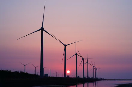 HD desktop wallpaper showing a row of man-made wind turbines silhouetted against a vibrant sunset sky over calm water.