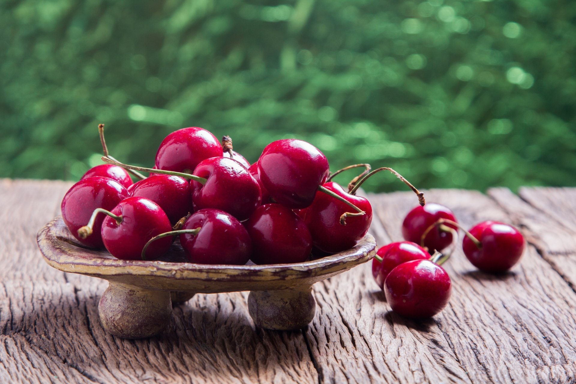 Glossy red cherries piled in a small pedestal bowl on rustic wood with blurred green bokeh — 4K Ultra HD PC desktop wallpaper and background (fruit, food, cherry).