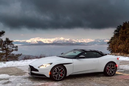 A white Aston Martin DB11 grand tourer parked on a snowy mountain road with a dramatic cloudy sky and snow-covered peaks in the background.