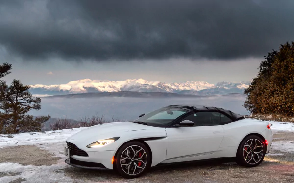 A white Aston Martin DB11 grand tourer parked on a snowy mountain road with a dramatic cloudy sky and snow-covered peaks in the background.