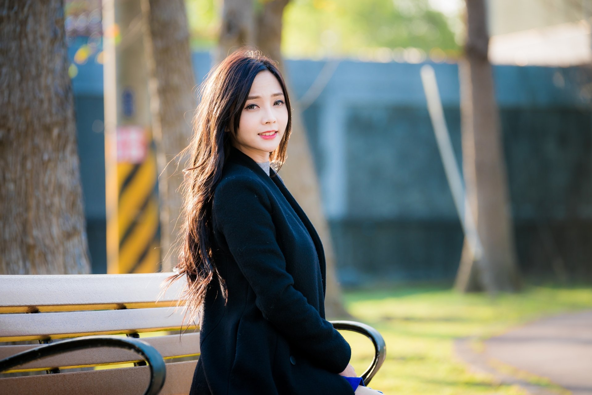 A smiling brunette woman with long hair and wearing a coat sits on a park bench. The background features a depth of field effect with trees and a blurred backdrop, creating a serene outdoor HD wallpaper.