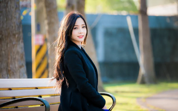 A smiling brunette woman with long hair and wearing a coat sits on a park bench. The background features a depth of field effect with trees and a blurred backdrop, creating a serene outdoor HD wallpaper.