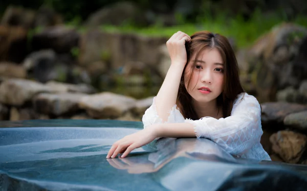 A high-definition desktop wallpaper featuring an Asian brunette woman with a serene expression, captured with a depth of field effect against a natural, rocky background.