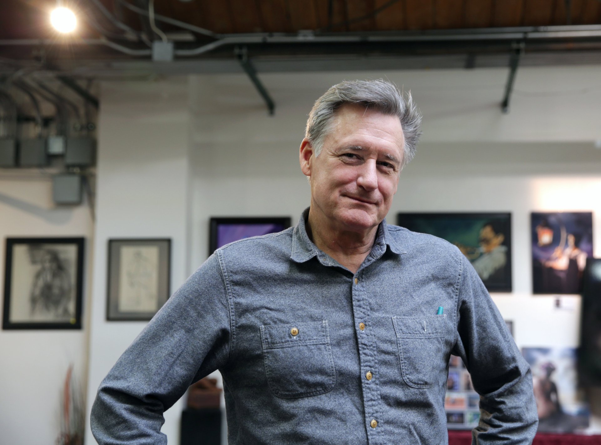 HD background featuring Bill Pullman smiling casually, with artwork displayed in the background, capturing a relaxed moment in an artistic setting.