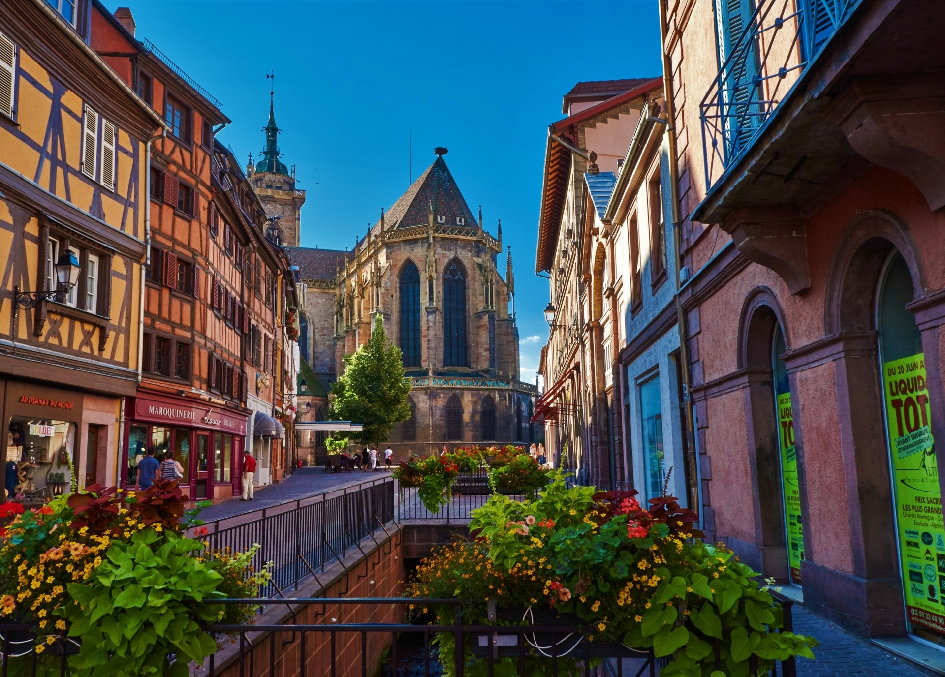 Colorful traditional buildings line a street in Colmar, France, with a historic church and vibrant flower boxes under a clear blue sky in this HD desktop wallpaper.